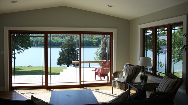 Une pièce dotée d’une grande porte-fenêtre coulissante qui donne sur une terrasse offrant une vue sur un lac et la verdure environnante. On y trouve un ventilateur de plafond, un canapé et une table avec des chaises.