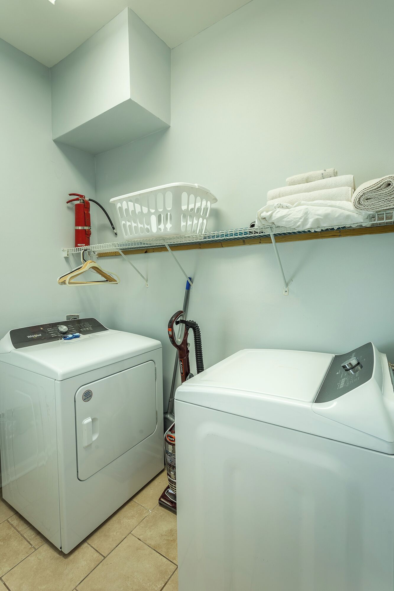 Une buanderie équipée d’un lave-linge à chargement frontal, d’un lave-linge et d’un sèche-linge superposés, d’un extincteur, d’un panier à linge et d’une étagère où sont pliées des serviettes.