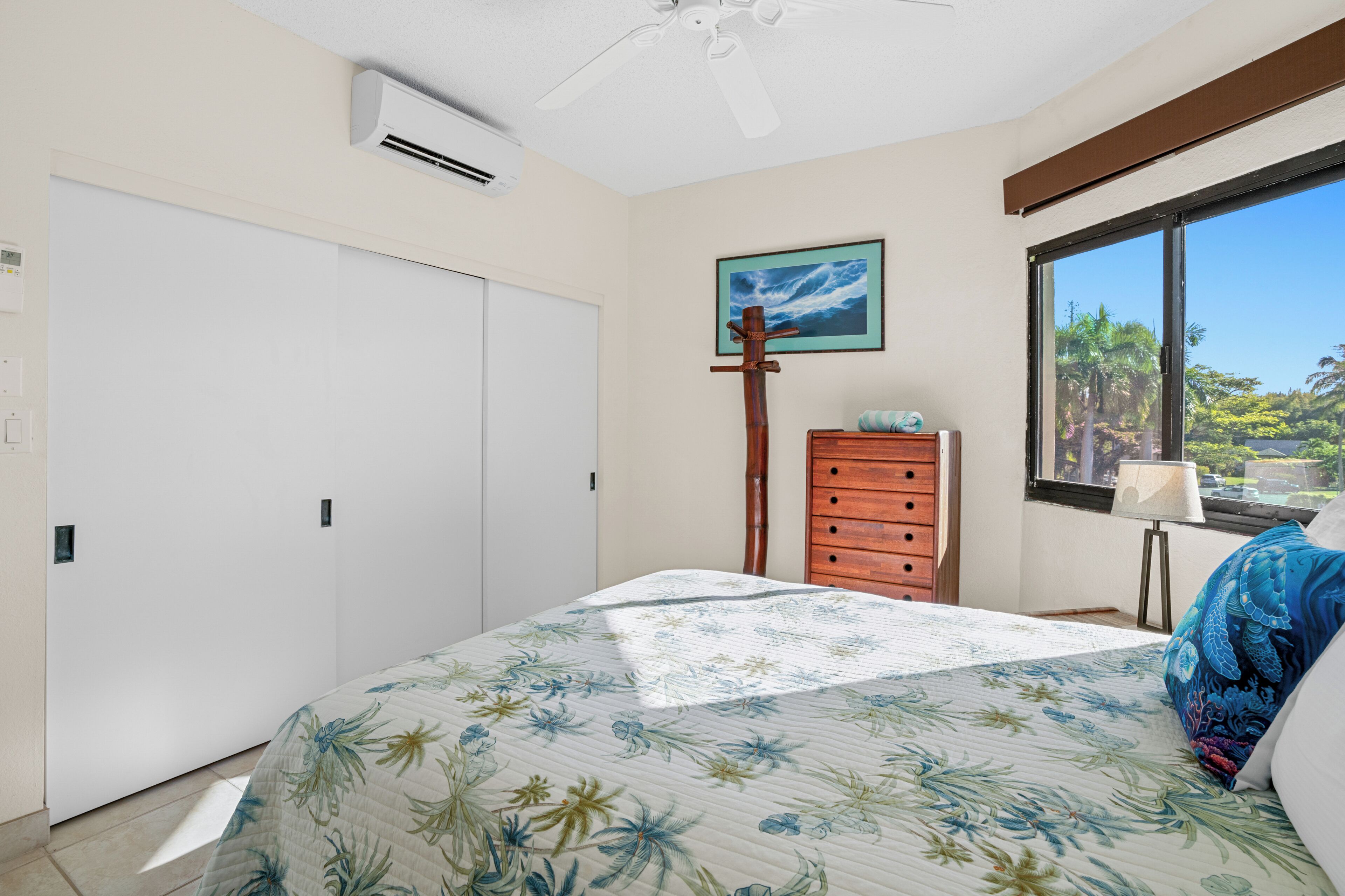 A bedroom with a bed, a dresser, a ceiling fan, an air conditioning unit, a window with a view, and a painting on the wall.