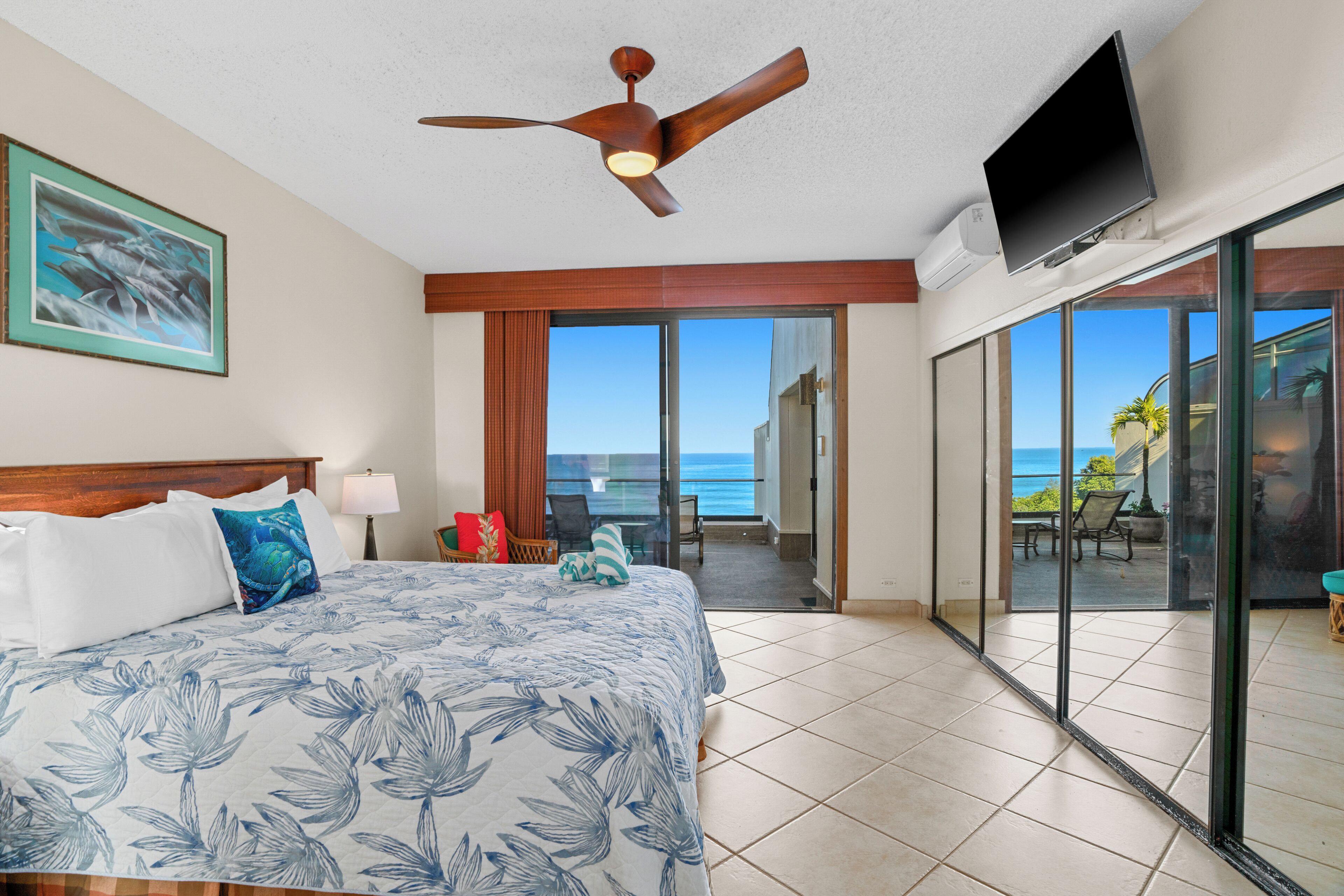 A hotel room with a bed, a ceiling fan, a TV, and a balcony with a view of the ocean.