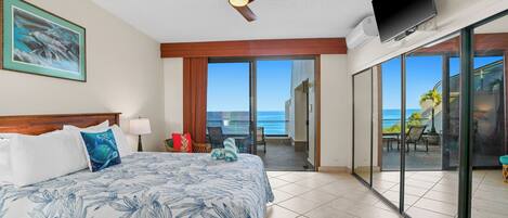 A hotel room with a bed, a ceiling fan, a TV, and a balcony with a view of the ocean.