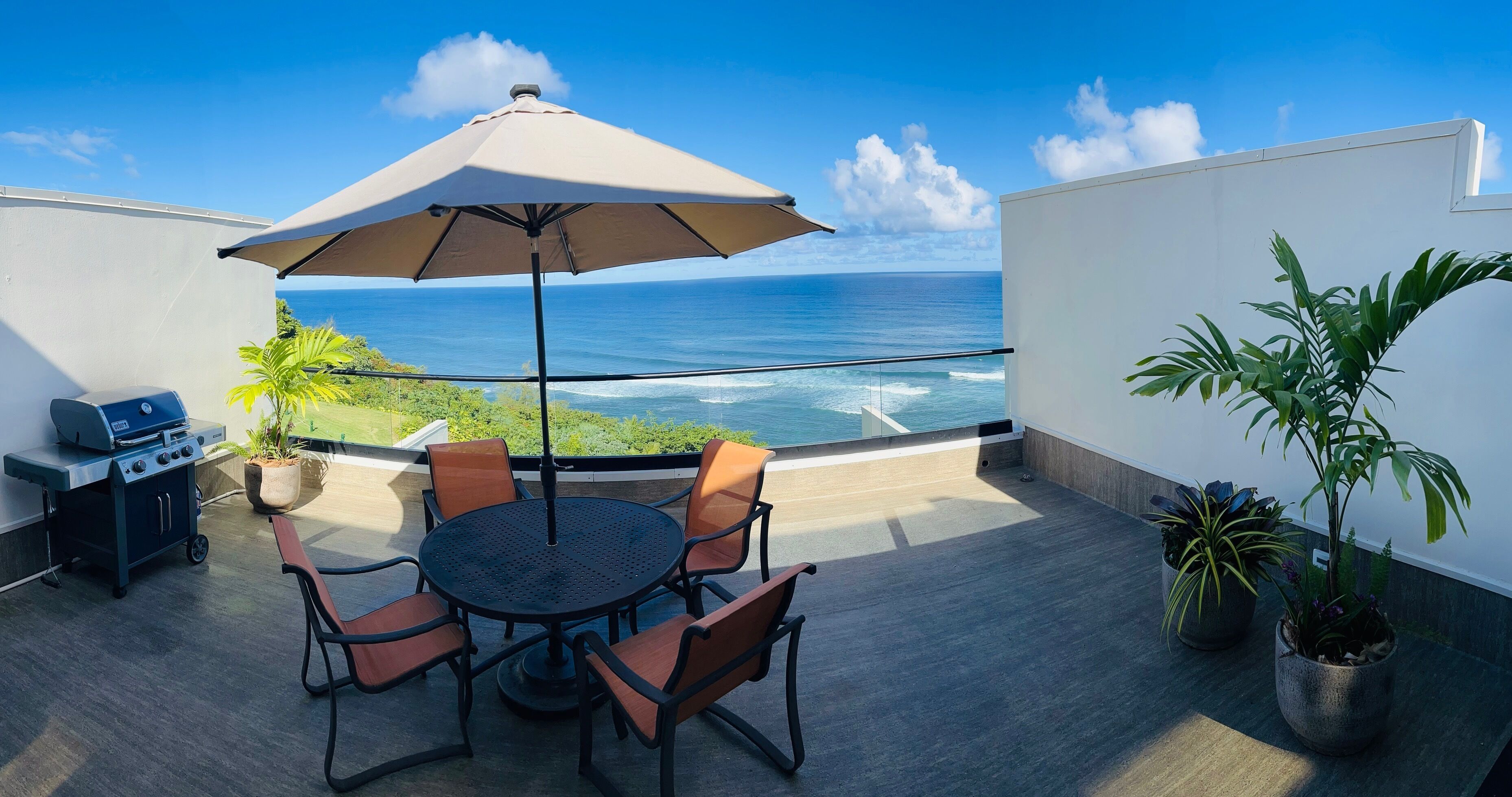 A rooftop terrace with a round table, chairs, a grill, and a large umbrella.