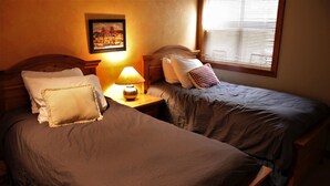 Une chambre avec deux lits simples, une table de chevet avec une lampe, un tableau encadré au mur et une fenêtre avec des stores.