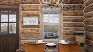 Ein rustikales Blockhaus-Interieur mit einem Esstisch für zwei Personen, einer Kandelaber und einem Fenster mit Blick auf die Berge.