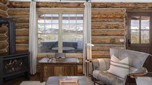 Ein Blockhaushaus-Interieur mit Kamin, einer Couch, einem Tisch und Blick auf schneebedeckte Berge durch ein großes Fenster.