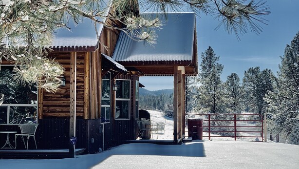 Una cabaña de madera con un porche cubierto, rodeada de árboles cubiertos de nieve y un cielo despejado.