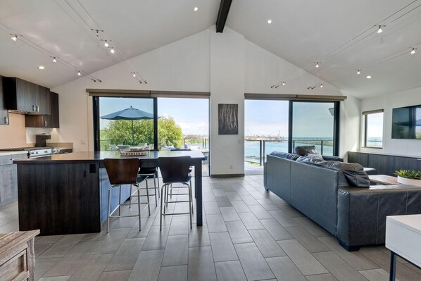 A spacious kitchen with a central island, bar stools, and a large dining table. There is a grey sofa and a TV mounted on the wall. The room has large windows offering a view of the sea.