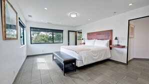 A modern bedroom with a large bed, a bench, and a view of greenery through the windows. - Kai Entire Home (5 bedroom / 5 bath) (Carlsbad)