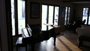 A spacious living room with a grand piano, large windows, and a view of a snowy landscape. - Cozy Lake Winnebago Cottage: Shallow Sandy Shore, Close to EAA & Packers Games (Menasha)