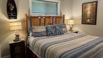 Un dormitorio con cabecera de madera, colcha a rayas, dos lámparas de noche, una mesita de noche, un cuadro en la pared y una ventana con persianas.