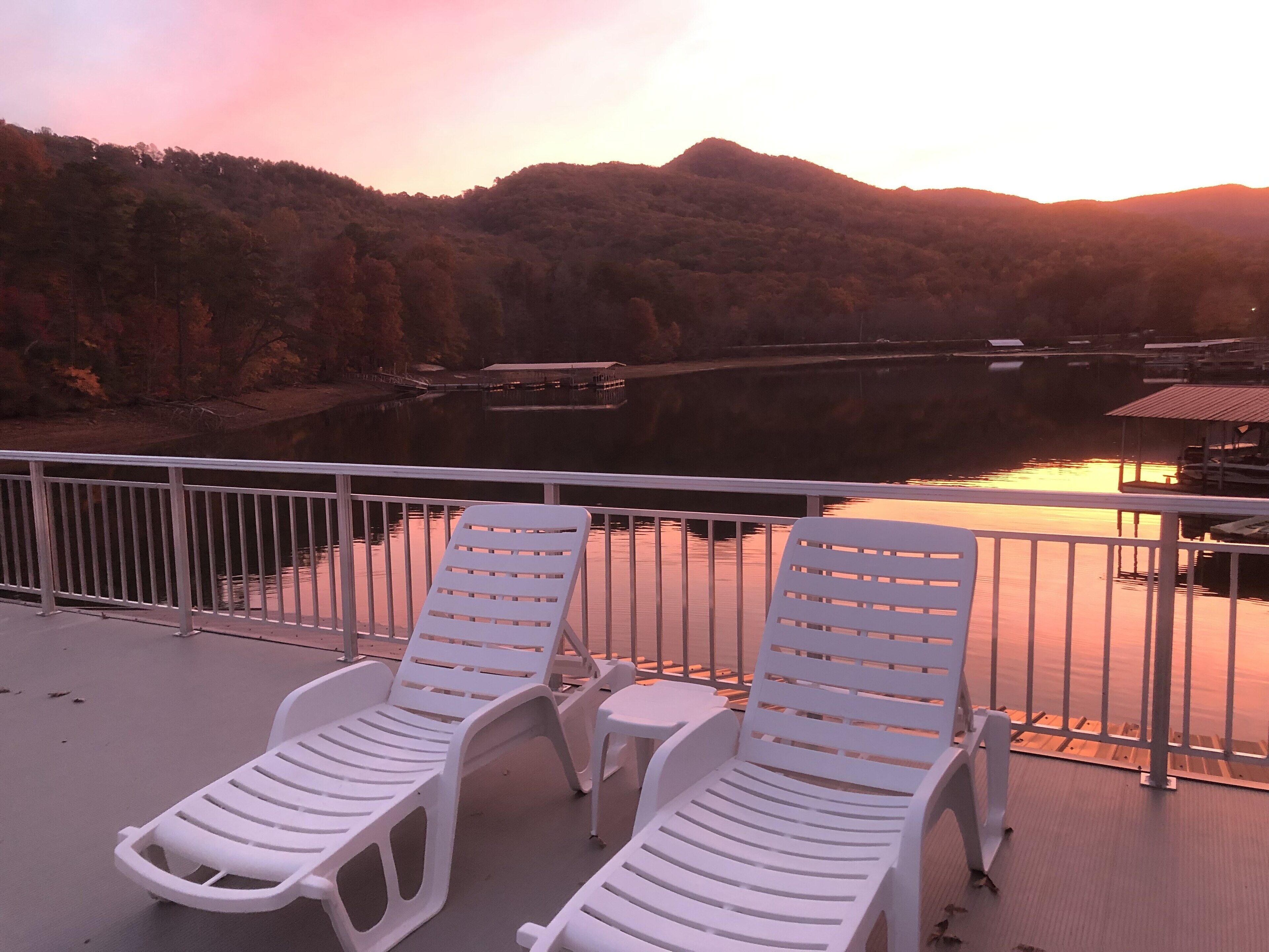 Deux fauteuils de jardin blancs sont placés sur une terrasse dotée d’une balustrade, d’où l’on peut admirer un plan d’eau paisible et un paysage vallonné au coucher du soleil.