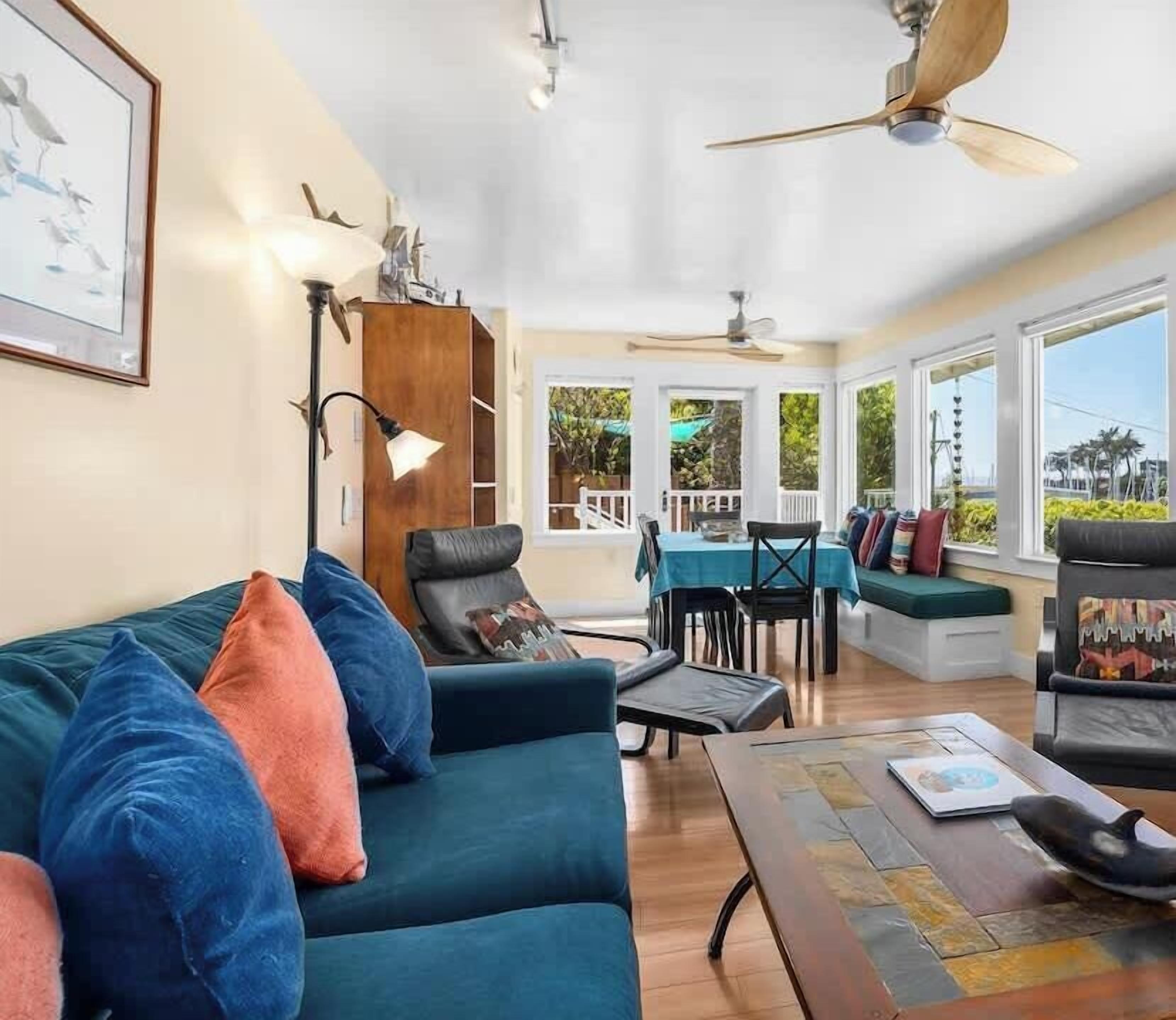 A spacious living room with a teal sofa, a dining table, and chairs. There is a ceiling fan, a floor lamp, and large windows offering a view of the outdoors.