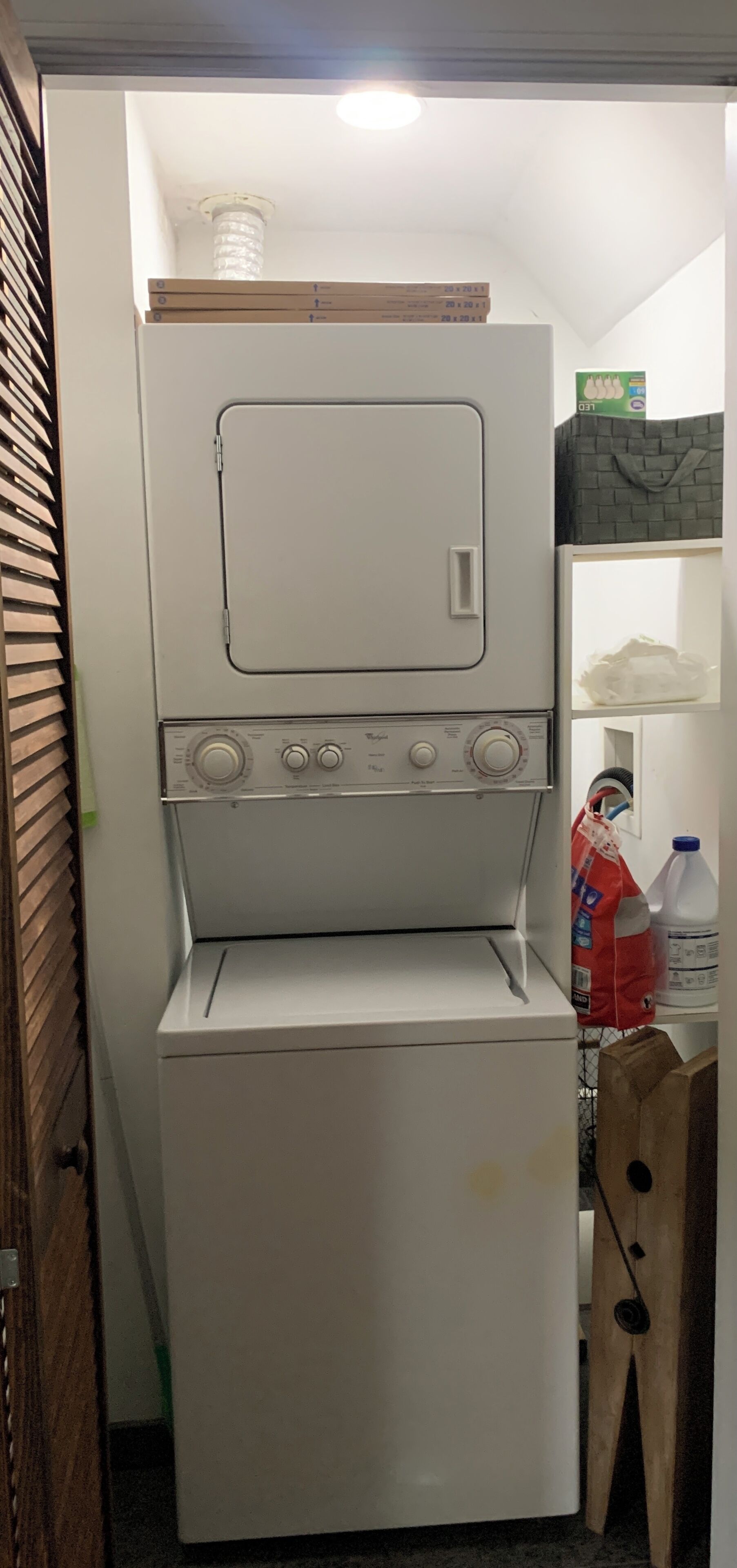 Een kleine, witte wasruimte met een inbouw wasmachine en droger, een houten plank boven de droger, en een klein opbergvak met een waterkan en een plastic zak.