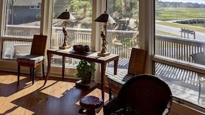 Quarto com janelas amplas, piso de madeira e uma mesa com cadeiras e luminárias.