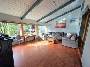 Un salon avec un plancher en bois, un canapé gris, une petite table et une chaise. Il y a des fenêtres qui laissent entrer la lumière naturelle et offrent une vue sur la verdure à l’extérieur.