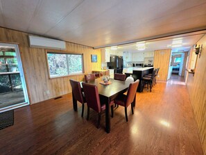 A spacious dining area with a wooden table and chairs, a kitchen in the background, and a view of the outdoors through a large window.