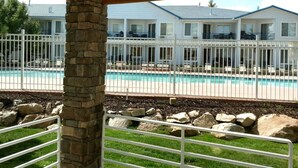 A pool area with a fenced deck, stone pillar, and a view of a row of white buildings with balconies.