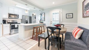 A compact kitchen with white cabinets, a central island, and a dining area with a gray sofa and wooden chairs. - Ocean View 2BR Condo Near Pier Village | St. Simons Island Vacation Rental (St Simons Island)