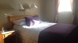 Una cama bien hecha con una colcha estampada y una almohada morada, una mesita de noche con una lámpara y un pequeño dispositivo electrónico, y un aplique de pared.
