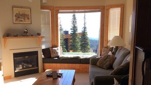 Una sala de estar acogedora con chimenea, un sofá, una mesa de centro y vistas a árboles a través de amplios ventanales.