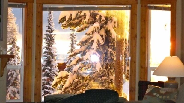 Una sala de estar acogedora con un amplio ventanal que muestra un paisaje invernal. Hay un sofá y una lámpara sobre una mesita auxiliar.