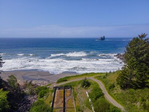 Un paysage côtier avec un promontoire rocheux qui s’avance dans la mer, une plage de sable et un sentier sinueux menant à une construction surmontée d’un réservoir métallique.