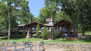 A lodge with a stone wall, a wooden deck, and a flag. - Beautiful 7 Bed, 6 Bath, Huge Dock, Boat Launch @ House, Game Room, Kid Friendly (Sunrise Beach)