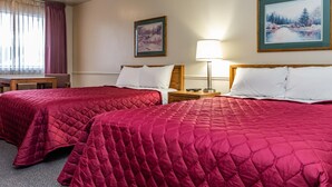 A hotel room with two beds, each with a red quilted bedspread and white pillows. There is a wooden nightstand with a lamp between the beds, and a painting of a winter scene above the nightstand. - Manti Country Village Motel (Manti)