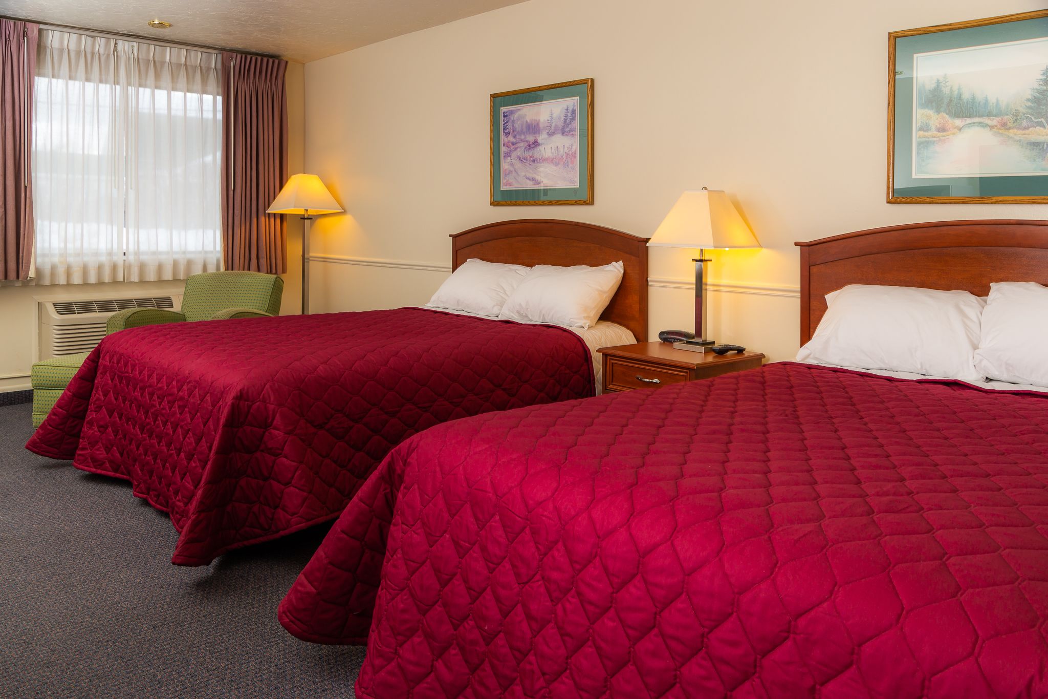 Ein Hotelzimmer mit zwei Betten, jeweils mit roter gesteppter Bettdecke, einem Holz-Kopfteil und einem Nachttisch mit Lampe. Es gibt ein Fenster mit Vorhängen, ein an der Wand hängendes Bild und einen grünen Sessel.