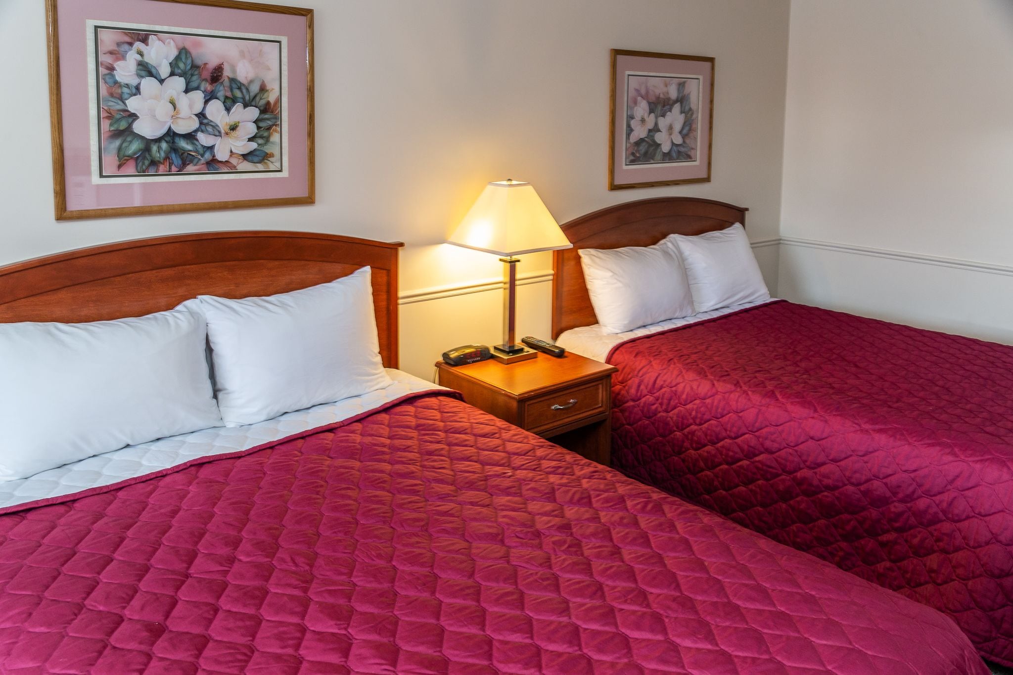 Een hotelkamer met twee bedden, elk met een rode geborduurde spreideken en witte kussens. Daartussen staat een nachtkastje met een lamp.