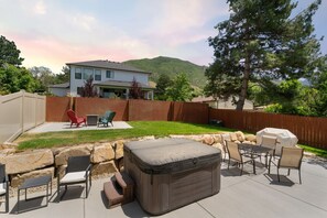 Une terrasse à l’arrière de la maison, aménagée avec du mobilier d’extérieur, un jacuzzi et offrant une vue sur une maison et des montagnes.