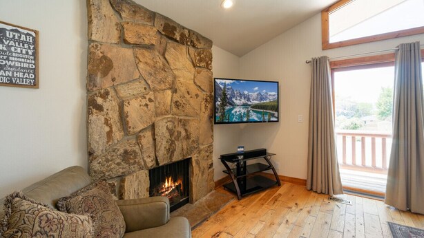 A cozy living room with a stone fireplace, a flat-screen TV mounted above it, and a large window with curtains. - Minutes to mountains resorts & downtown SLC (cottonwood heights)