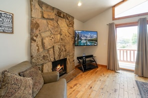 A cozy living room with a stone fireplace, a flat-screen TV mounted above it, and a large window with curtains. - Minutes to mountains resorts & downtown SLC (cottonwood heights)