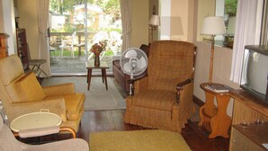 A living room with a sofa, armchair, and a television. There is a fan, a small table with a vase, and a view of the outdoors through a glass door. - Bemus Point Vacation Rental (Bemus Point)