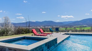 Une piscine entourée d’une terrasse en pierre, avec des chaises longues rouges et bleues, d’où l’on peut admirer des montagnes et des champs en arrière-plan.