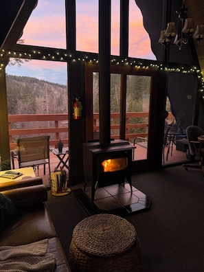 A cozy room with a wood-burning stove, a view of the forest, and a balcony with string lights.