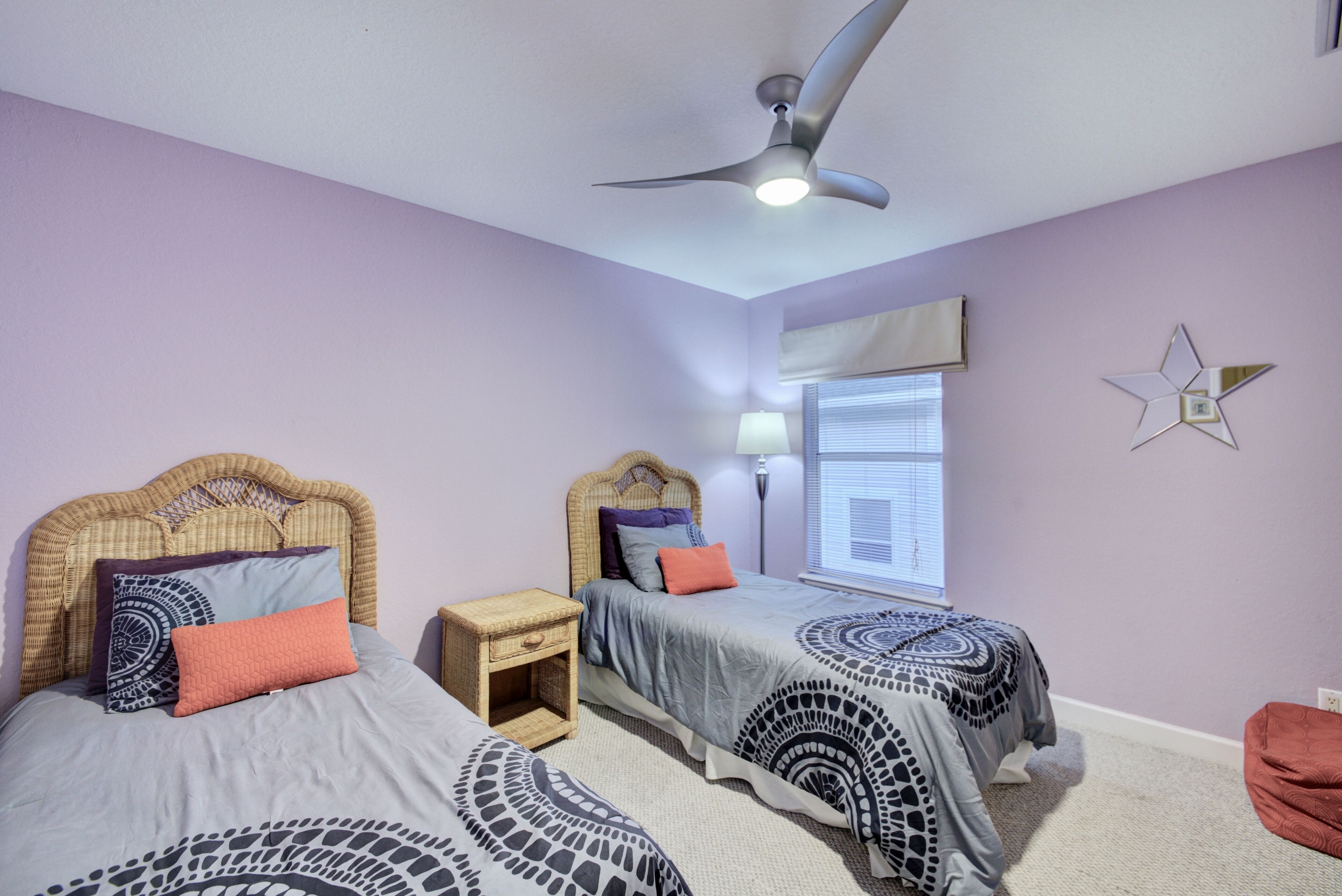 A bedroom with two beds, a ceiling fan, a bedside table, a lamp, a window with blinds, and a star decoration on the wall.