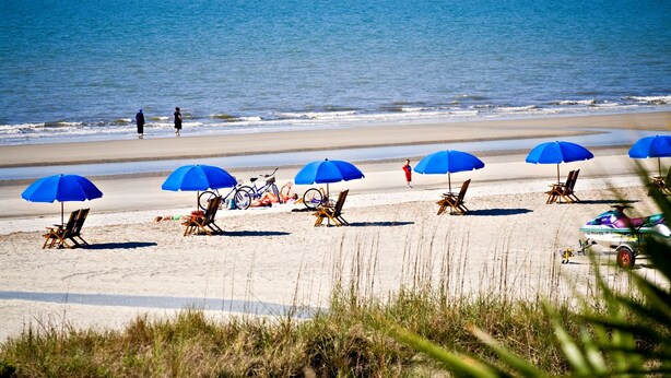 A beach with blue umbrellas, beach chairs, and a bicycle. - 121 Greens-7 Min Walk to Beach, Full Remodel w/Beach Locker & Chairs (Hilton Head Island)