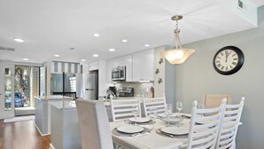 A modern kitchen with white cabinetry, a central island, and a dining area with white chairs and a table set for four. - 121 Greens-7 Min Walk to Beach, Full Remodel w/Beach Locker & Chairs (Hilton Head Island)