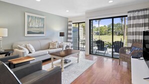 A modern living room with a sofa, coffee table, and chairs. There is a painting on the wall and a view of the outdoors through large windows. - 121 Greens-7 Min Walk to Beach, Full Remodel w/Beach Locker & Chairs (Hilton Head Island)
