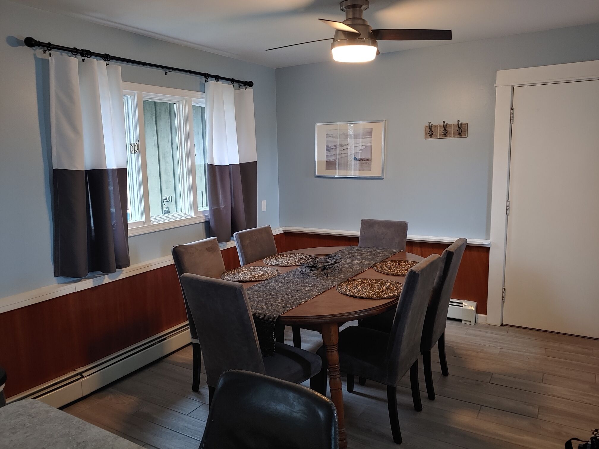 A dining room with a wooden table, six chairs, a ceiling fan, and a window with curtains.