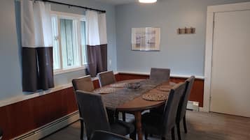 A dining room with a wooden table, six chairs, a ceiling fan, and a window with curtains.