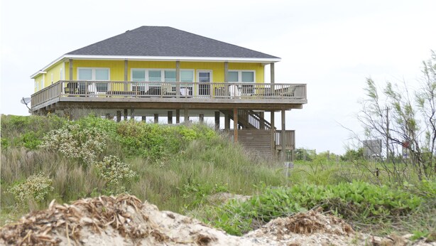 Ein zweistöckiges Haus mit einem Balkon im oberen Geschoss, umgeben von Vegetation und Sand.