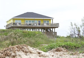 A two-story house with a balcony on the upper floor, surrounded by vegetation and sand. - Barney's Beach House - Ocean Front Fenced in yard for dogs (Gilchrist)