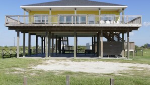 Ein Strandhaus mit zwei Etagen, einem Balkon, einer überdachten Terrasse und einer Treppe, die zum oberen Stockwerk führt.