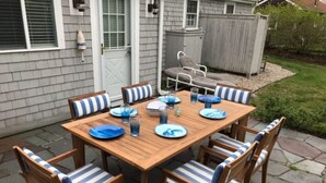Une table de patio en bois, entourée de chaises à rayures bleues et blanches, un petit barbecue et un ensemble de vaisselle comprenant des assiettes et des verres.