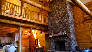 Interior de una cabaña de troncos con chimenea de piedra, muebles de madera y un balcón.