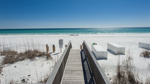 Ein hölzerner Steg führt zu einem Strand mit weißem Sand und klarem, blauem Wasser.