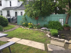 A backyard with a green fence, a garden with various plants and flowers, a stone pathway, and a metal table.