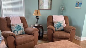A living room with two brown armchairs, a matching ottoman, a patterned rug, a lamp, and a framed artwork on the wall.